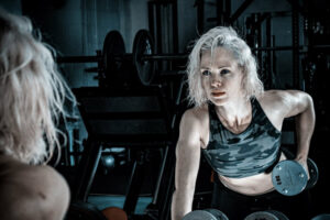 Woman in a dark gym stares intensely into mirror while lifting weights.