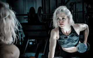 Woman in a dark gym stares intensely into mirror while lifting weights.