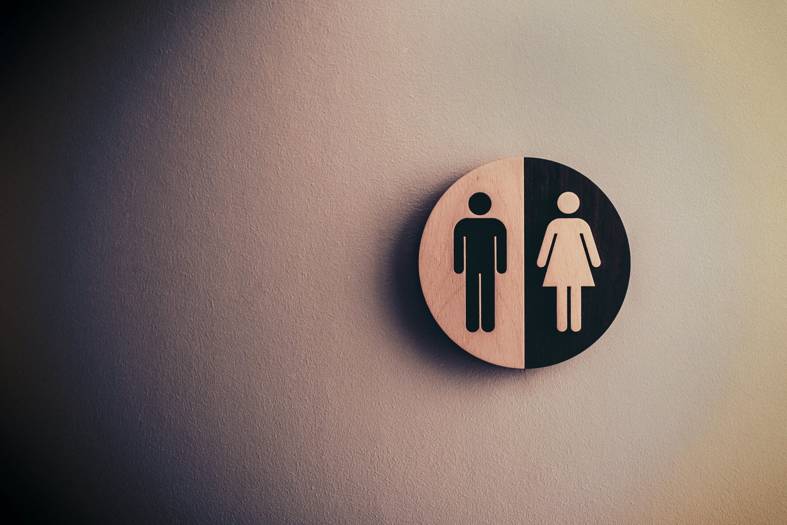 A circular wooden bathroom sign with a black-and-white motif depicting a male on the left half and female on the right half.
