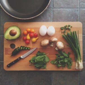 Wooden cutting board with a knife, green vegetables, mushrooms, an avocado, eggs, and cherry tomatoes on top.